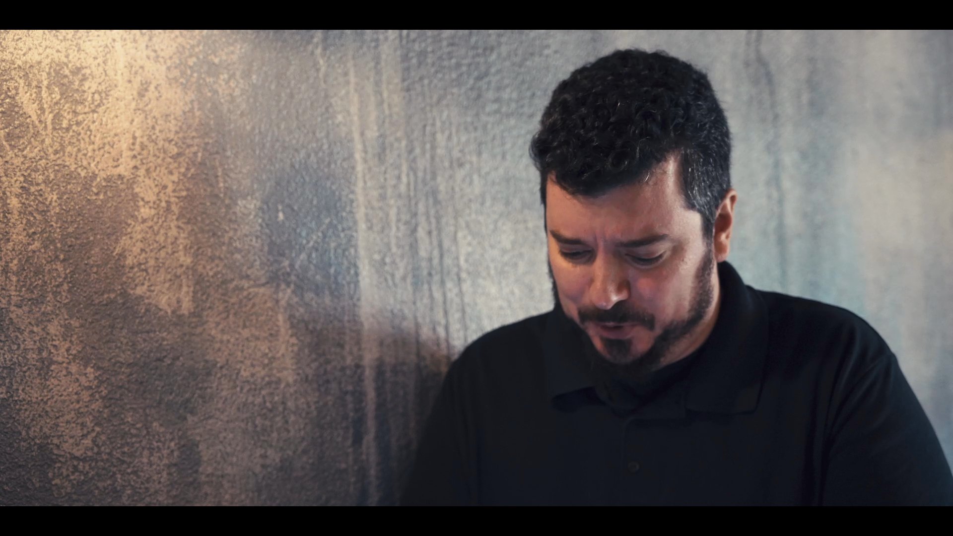 Two men engage in an earnest discussion, alternating speaking turns while seated against a textured wall.