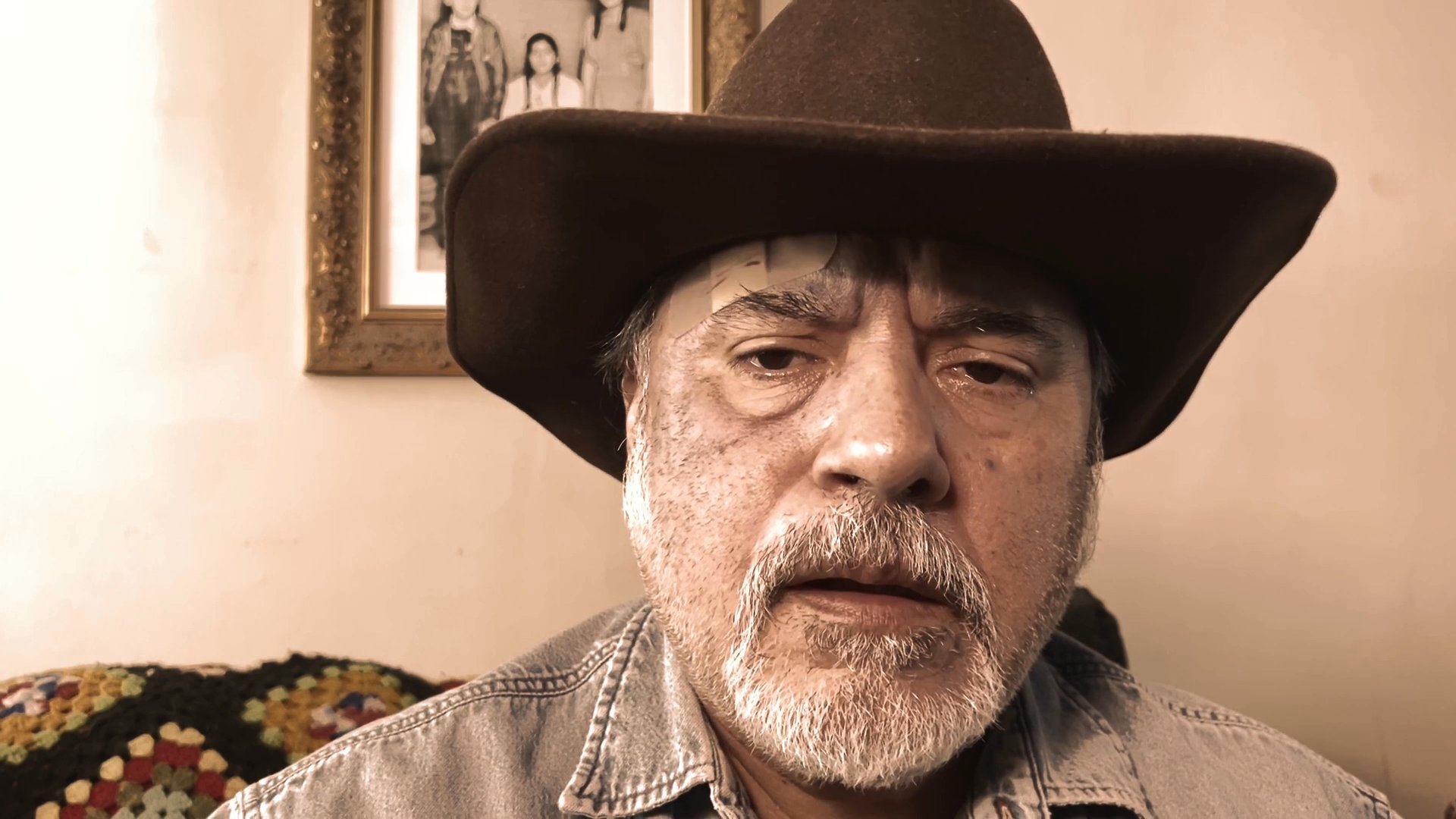 An older man wearing a brown cowboy hat speaks directly to the camera, conveying a narrative or commentary in a domestic setting.