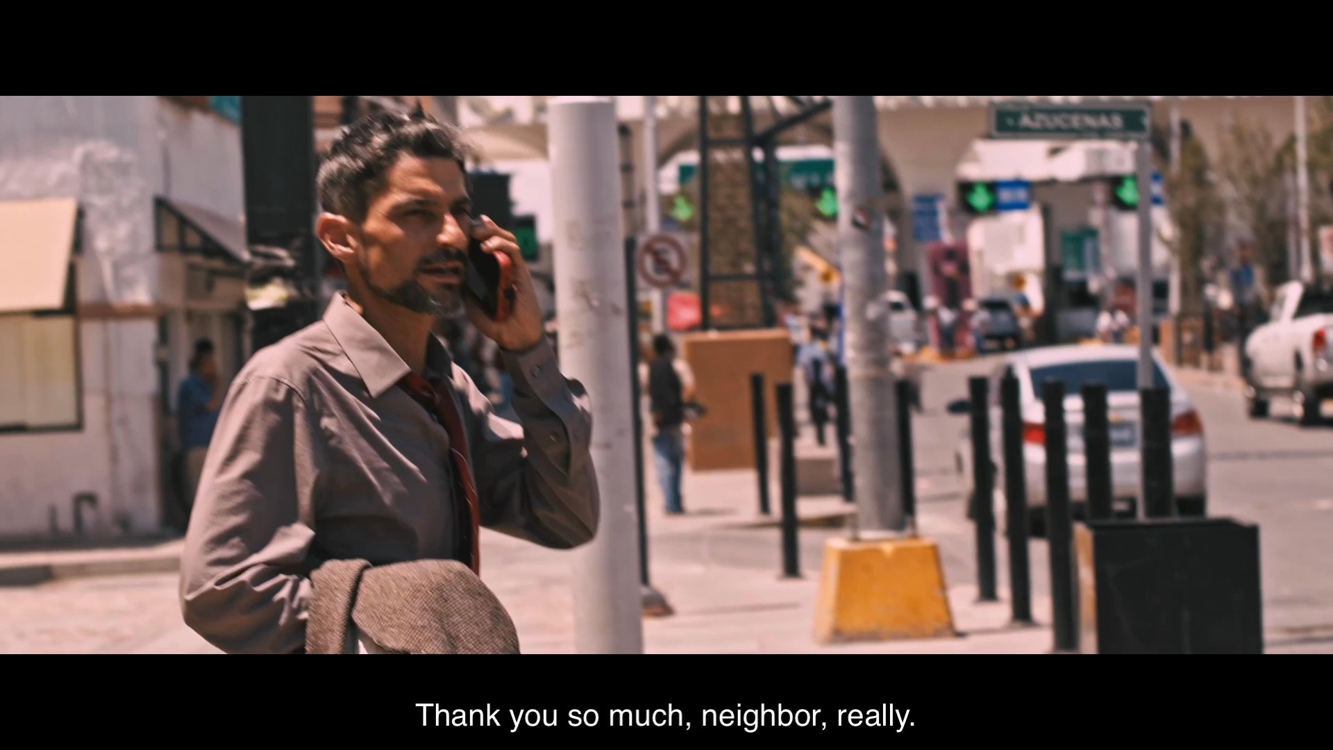 A man stands on a busy street corner while having a phone conversation, expressing gratitude and confirming plans to travel to a destination. The setting appears to be a public area near a road intersection with vehicle traffic.
