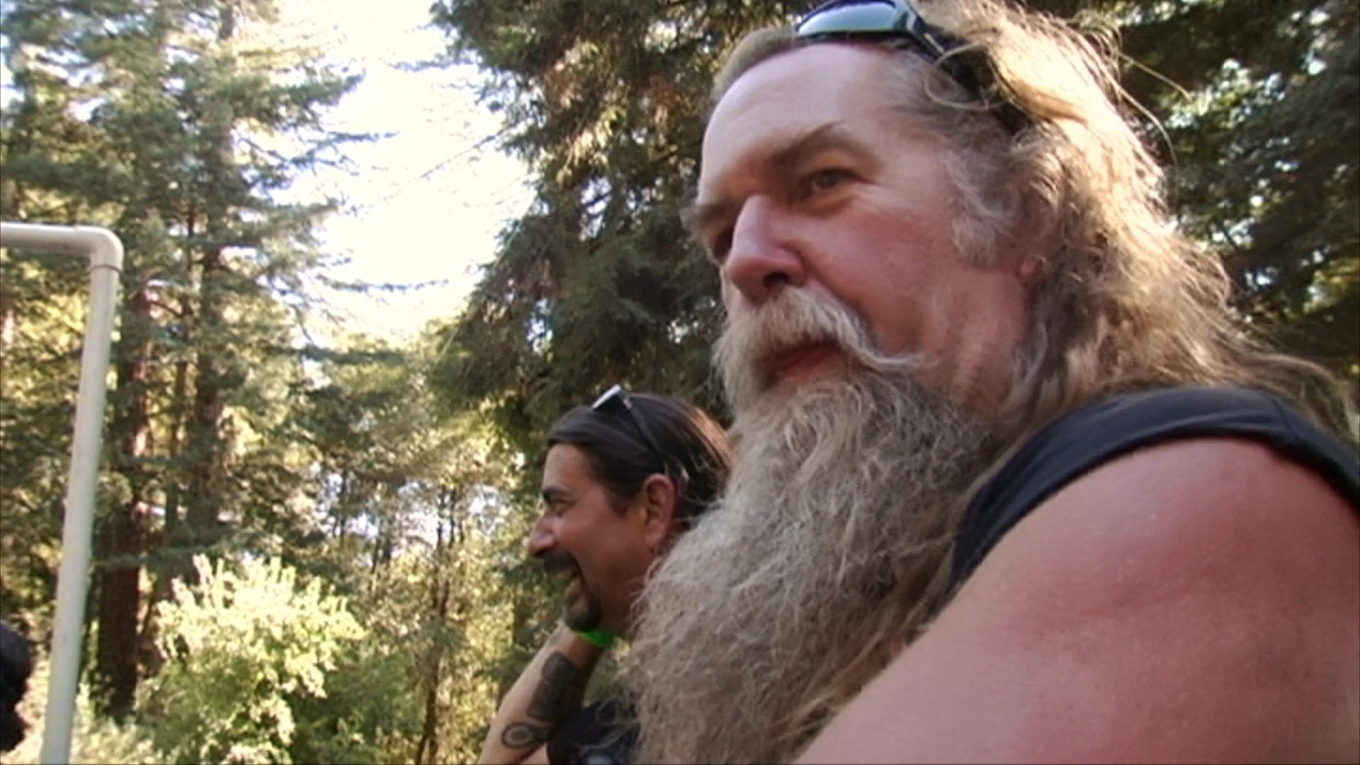 A man in a baseball cap gestures while speaking outdoors, followed by scenes of a motorcycle rally featuring club members and enthusiasts in a woodland setting.