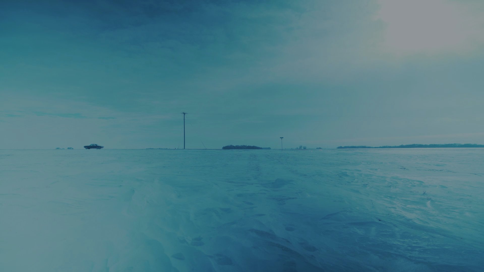 A vehicle traverses a desolate, snow-covered road spanning a vast, frozen landscape during a winter storm.