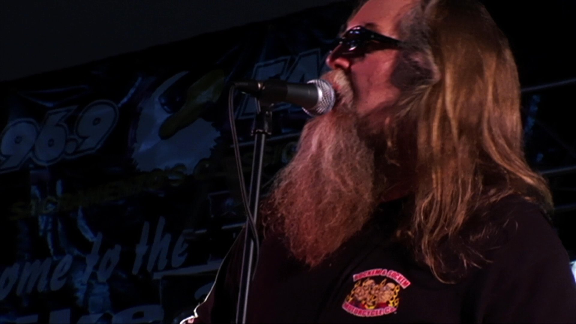 A bearded lead singer with long hair performs on stage with his rock band. The group plays instruments under professional stage lighting during a concert event.