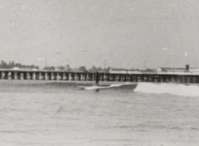 An elderly man discusses his past as a surfer while historical black-and-white footage of a man riding waves near a pier plays alongside him. The segments alternate between his current-day narration and vintage captures of the beach experience.