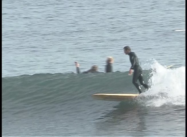 A surfer in a wetsuit maneuvers a surfboard across breaking ocean waves while other surfers wait in the distance. The individual maintains balance and control while navigating the swell near the coastline.