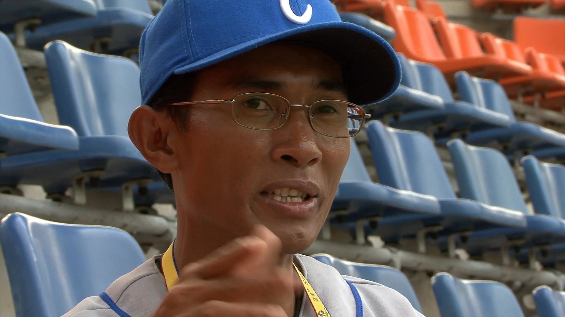 A man wearing a baseball cap and glasses delivers a commentary while seated in the empty bleachers of a sports stadium.