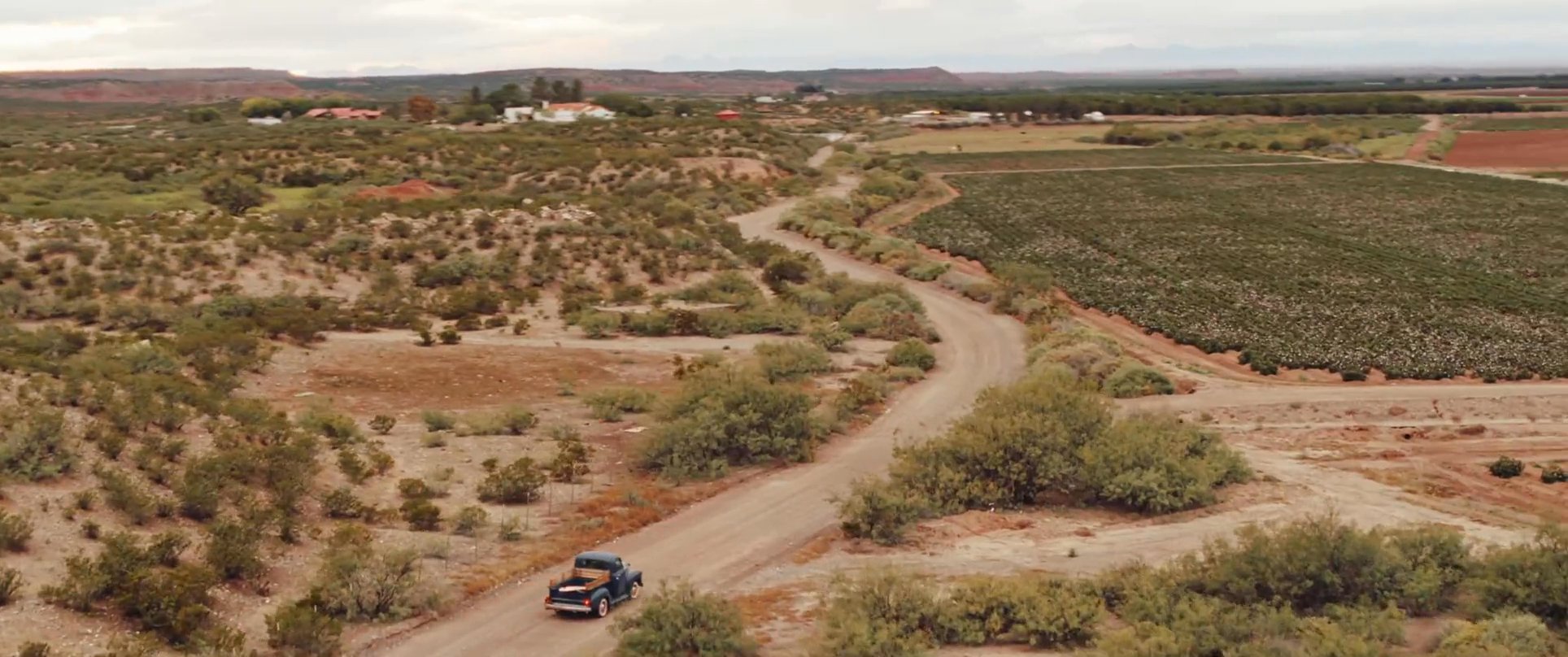 A man drives a vintage pickup truck through a desert environment and along rural roads surrounded by fields and orchards.