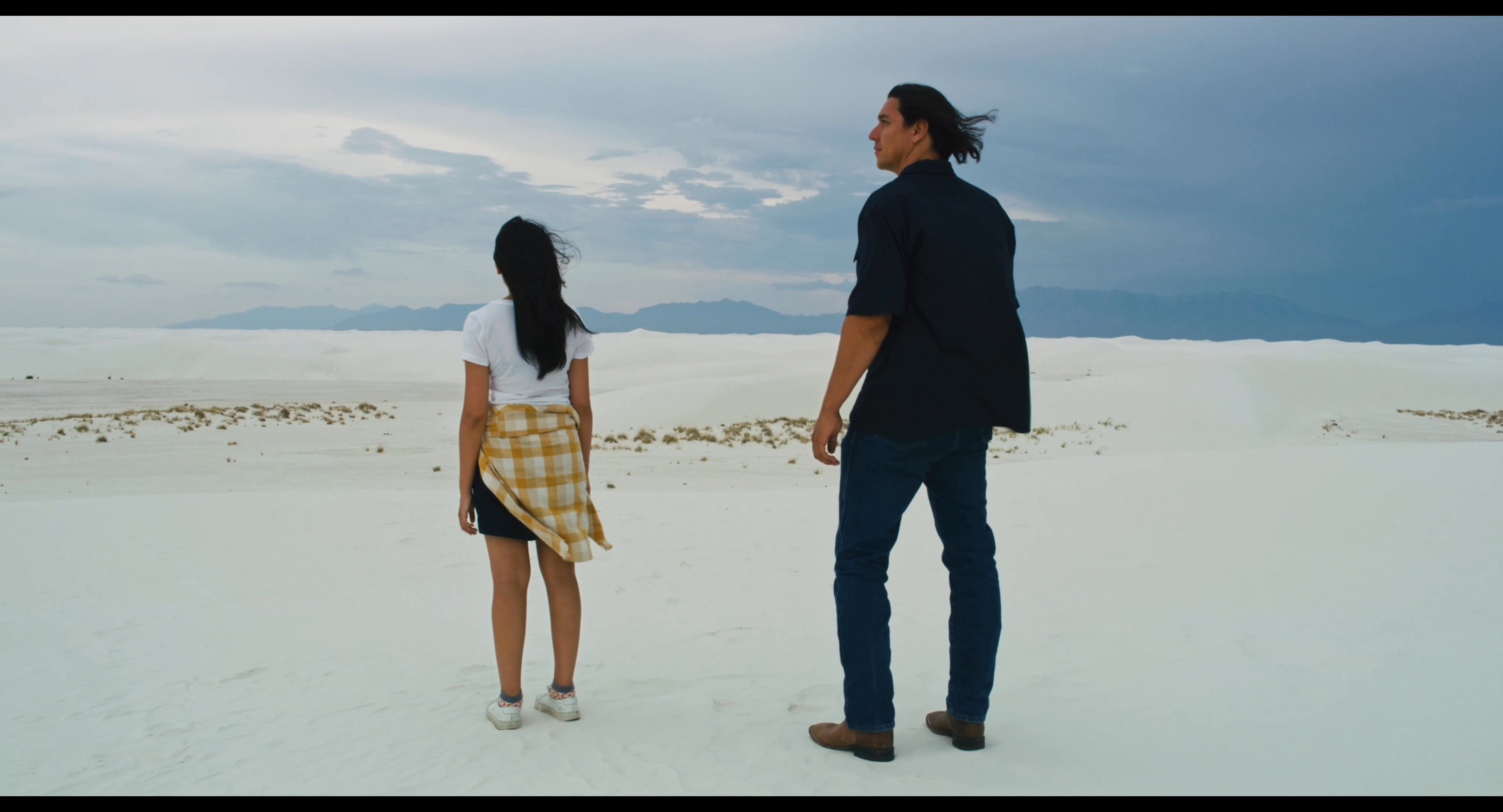 A man and woman walk together across a vast white sand dune landscape under an overcast sky. They observe their surroundings and enjoy a peaceful stroll through the remote terrain.