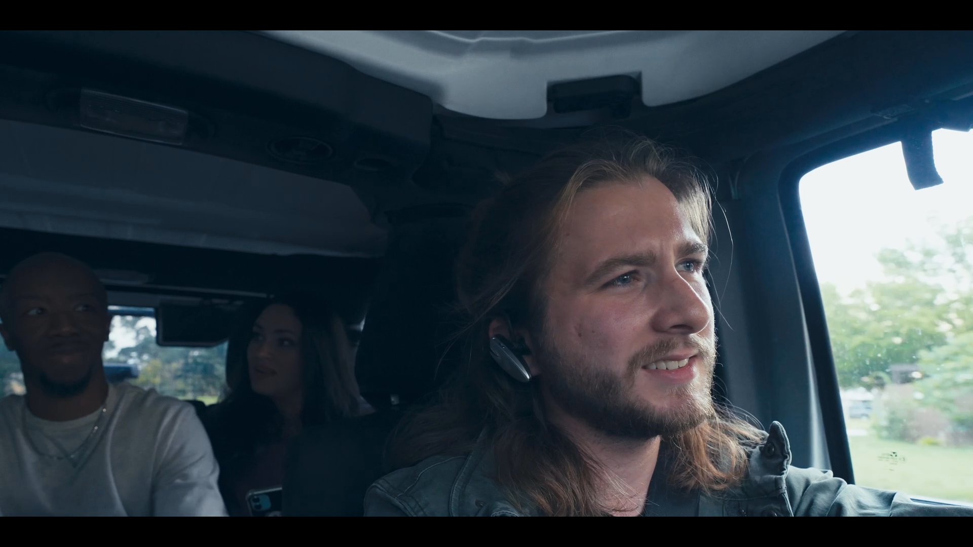 A man with long hair drives a vehicle while engaging in a casual conversation with passengers seated in the back.