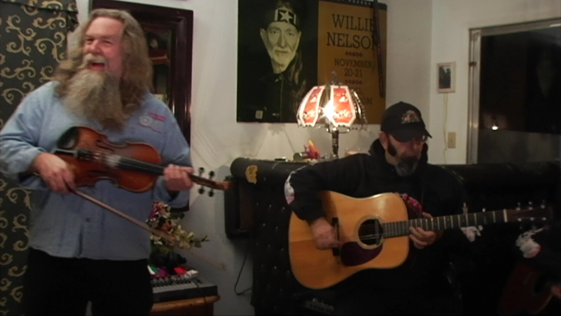 Two musicians perform a folk song in an indoor setting, with one individual playing an acoustic guitar and another playing the fiddle.