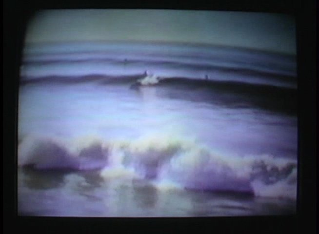 An elderly man shares historical context alongside archival footage of a surfer riding waves in the ocean. The scene highlights the evolution of surfing through a personal retrospective perspective.