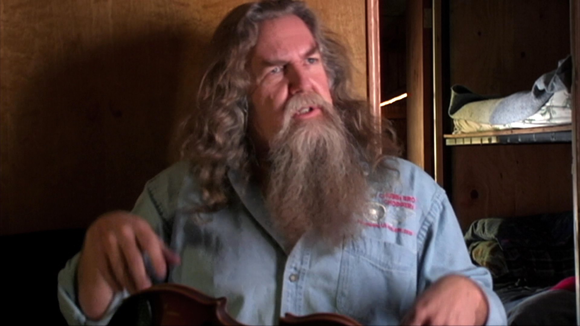 A man with long hair and a beard sits in a wood-paneled room, actively gesturing to emphasize his points during an instructional demonstration.