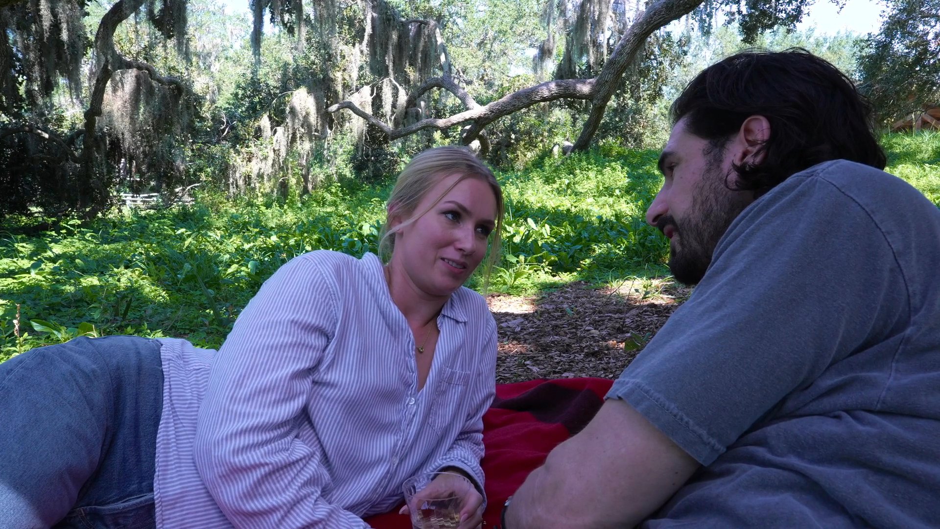 A man and a woman engage in conversation while reclining on a picnic blanket in a wooded, outdoor setting. The atmosphere is relaxed and intimate as they share a moment of leisure together.