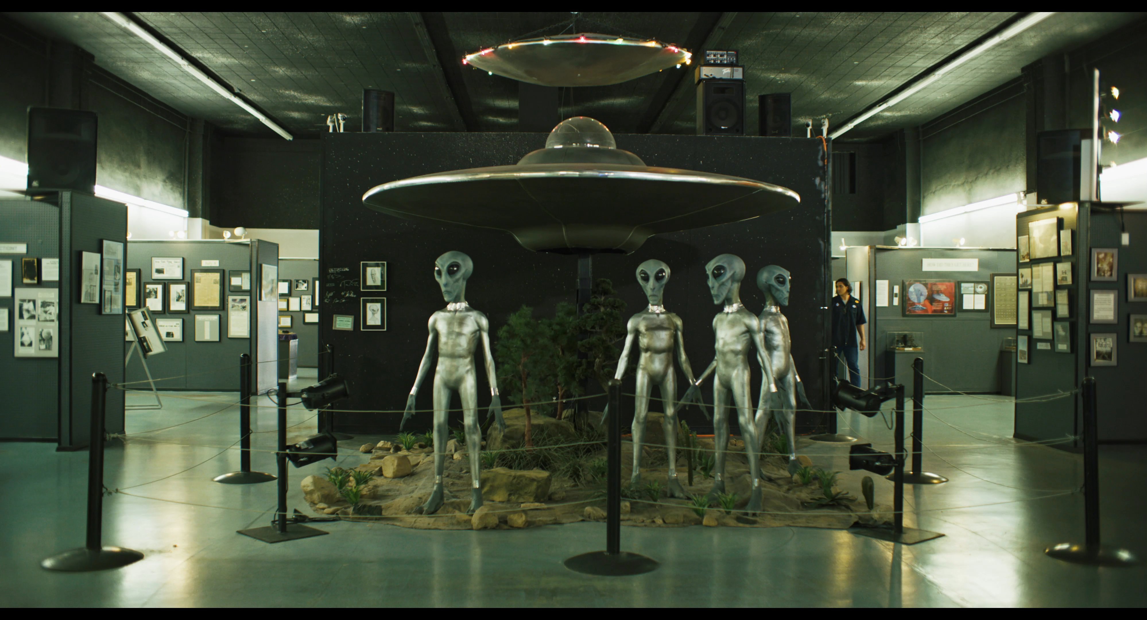Two individuals explore an extraterrestrial museum filled with displays of spacecraft models and alien figures.