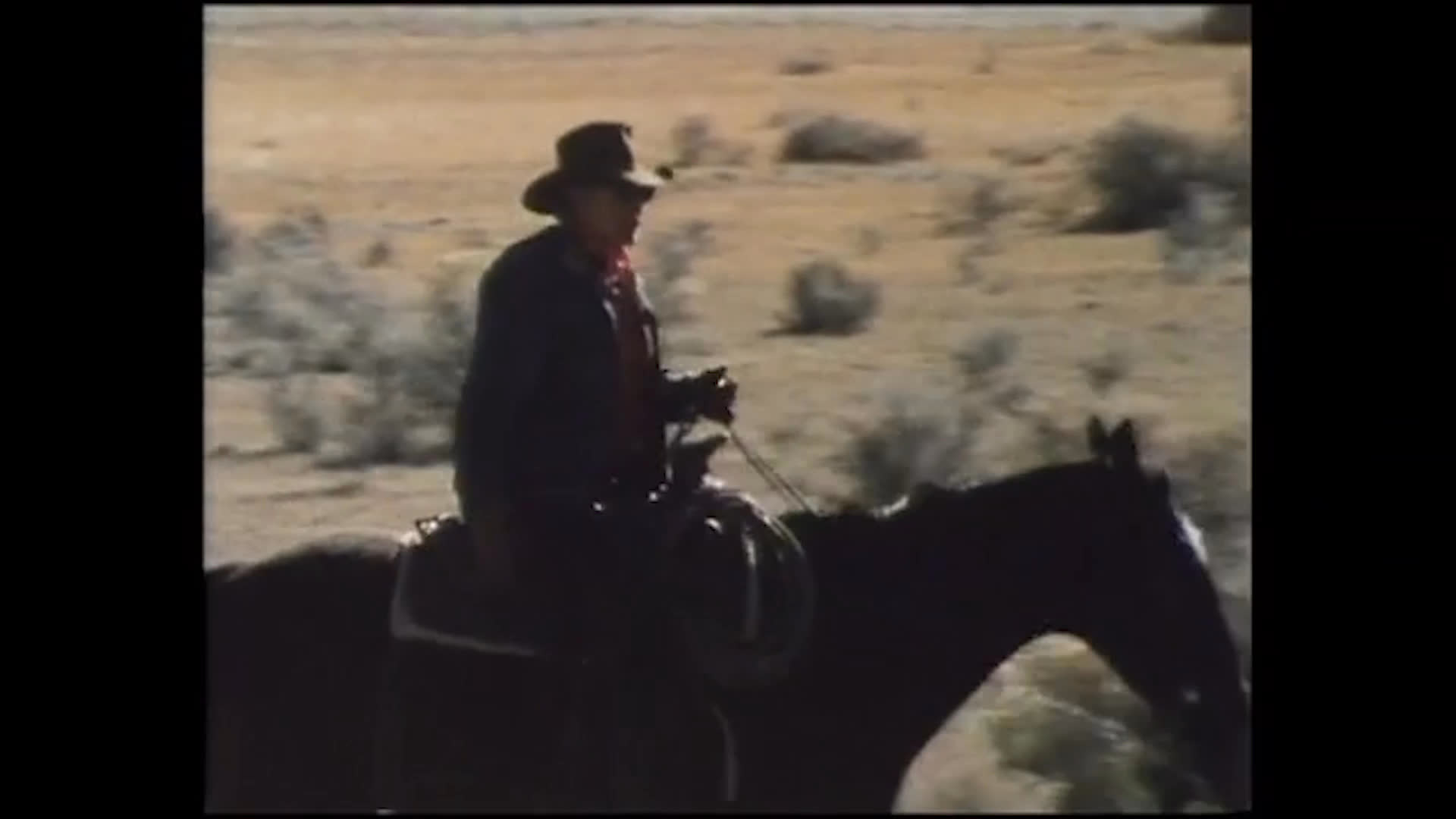 A cowboy rides across a dry, brush-covered range, with a herd of cattle in the distance. He smiles as he reflects on enjoying ranch work and says he would not like dressing up to go to town every day.
