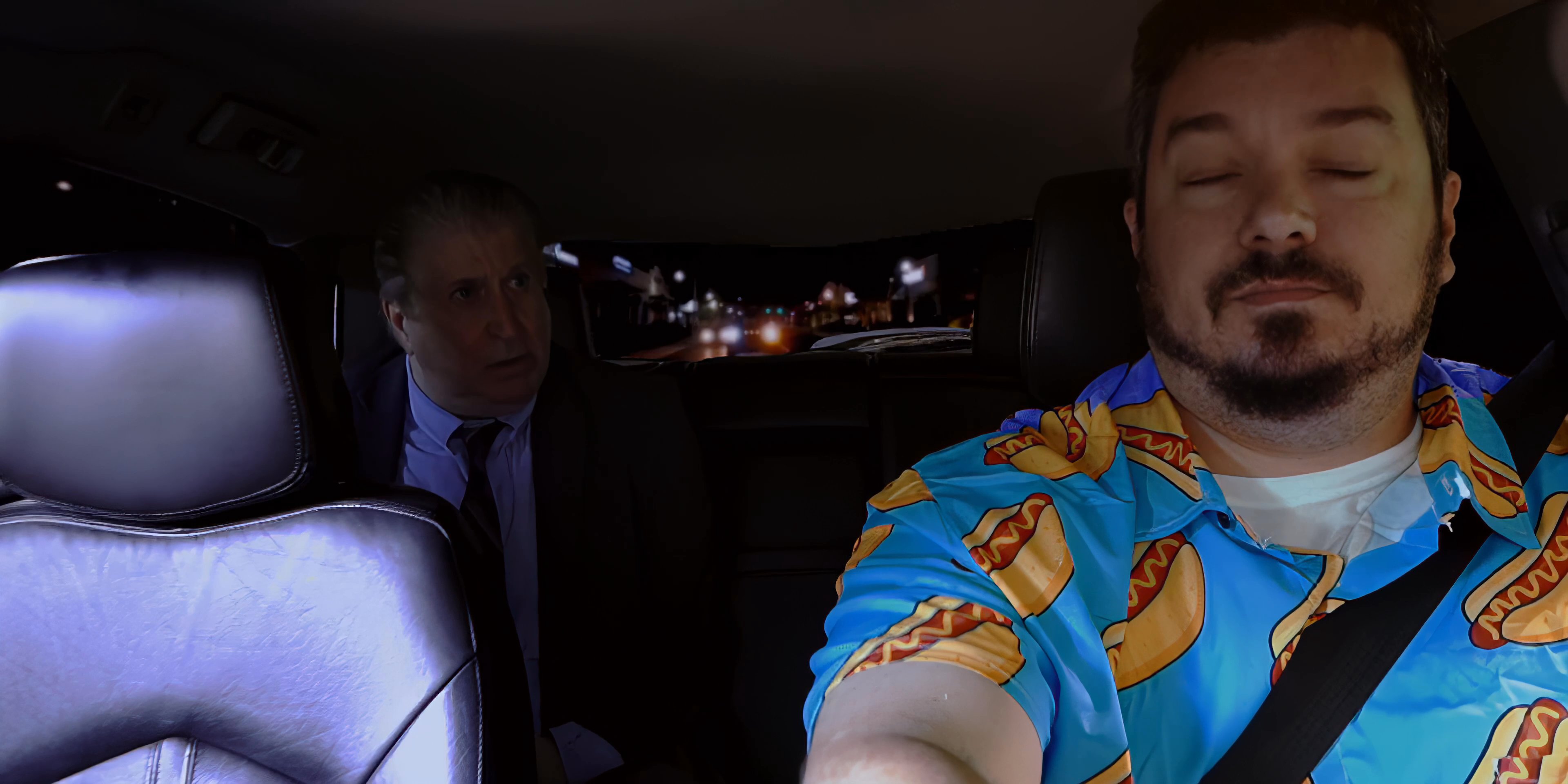 A man in a casual patterned shirt drives a passenger through a brightly lit urban environment at night. The two engage in a continuous conversation during the commute.