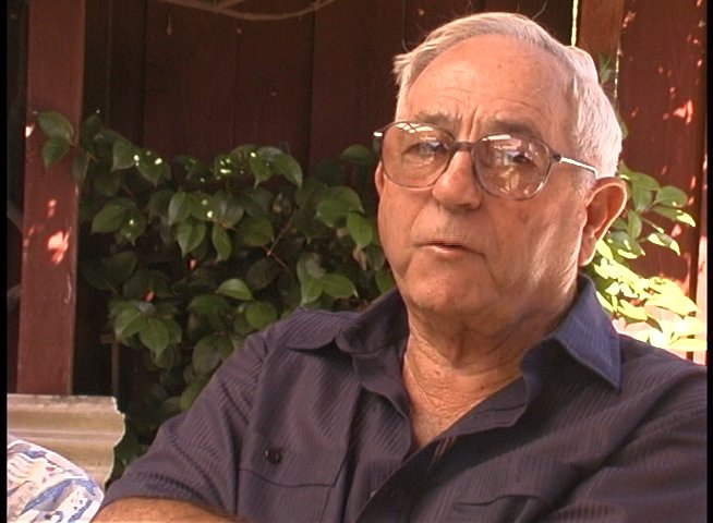 An older man with glasses sits outdoors and shares personal reflections or testimony during a formal interview.