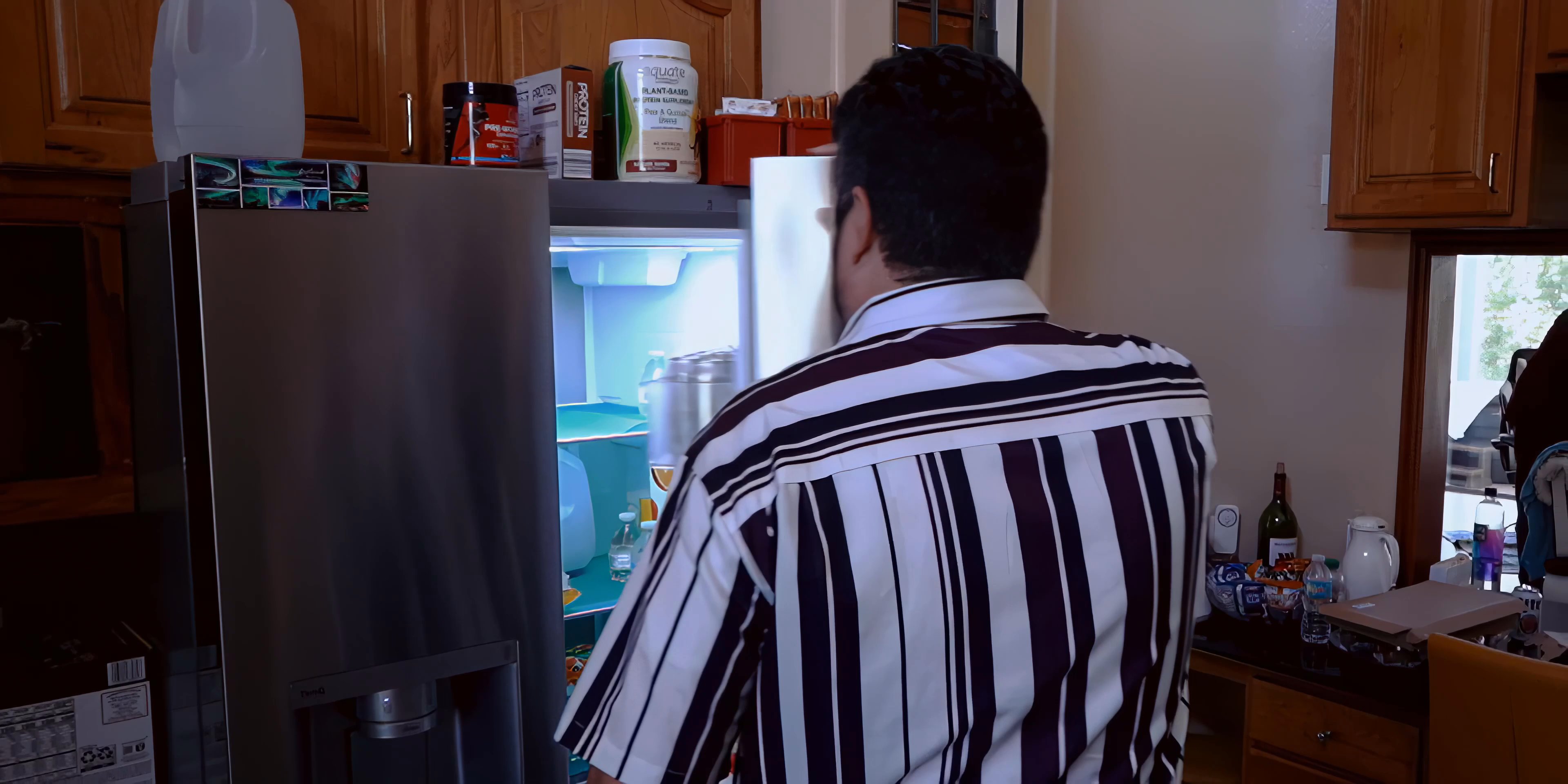 A man in a striped shirt accesses a refrigerator in a residential kitchen. The scene highlights standard household routines and kitchen organization.