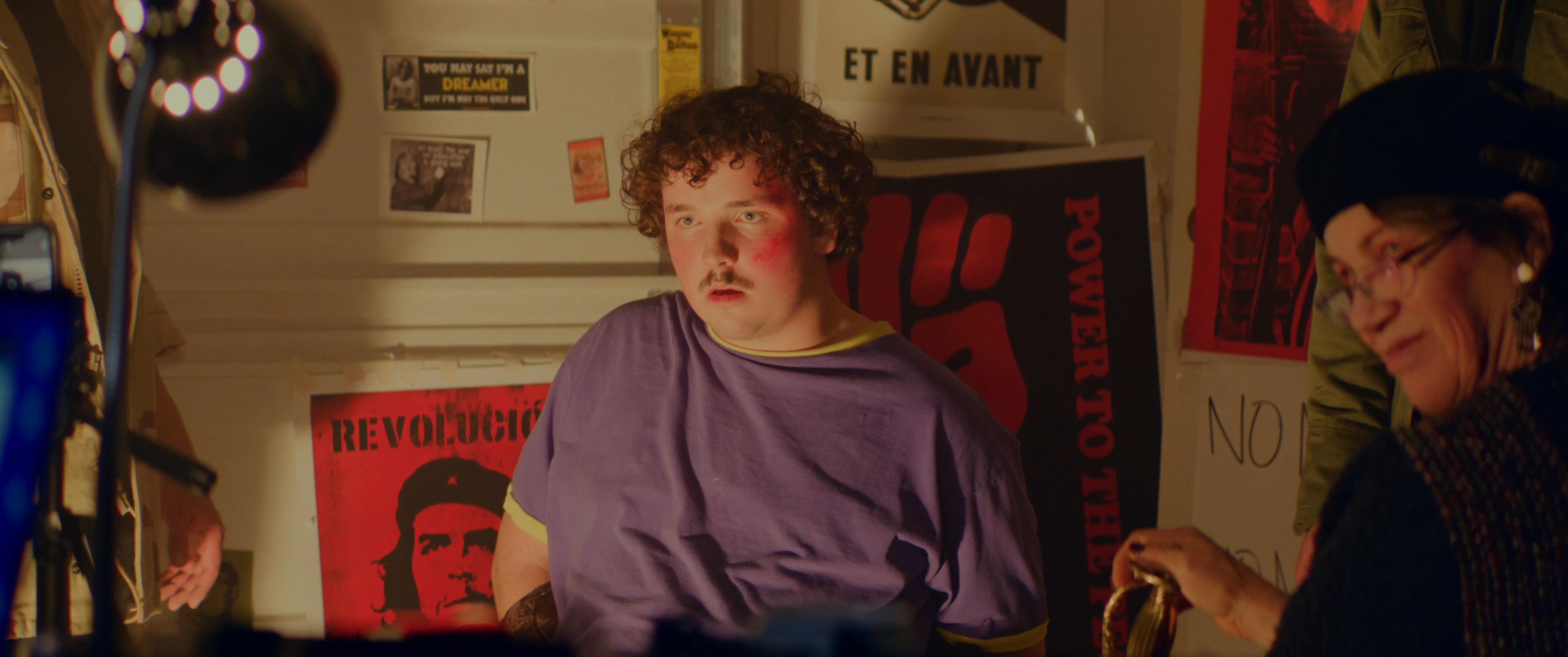 A diverse group of individuals collaborate in a workspace decorated with political posters and revolutionary imagery. The scene depicts a focused discussion regarding creative or organizational efforts.