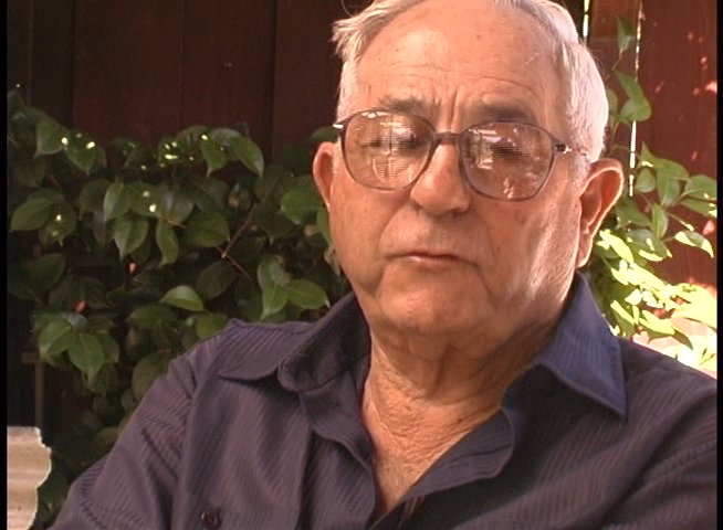 An elderly man provides personal testimony while reflecting on historical images displayed as visual overlays. He shares firsthand experiences and memories during a structured interview setting.