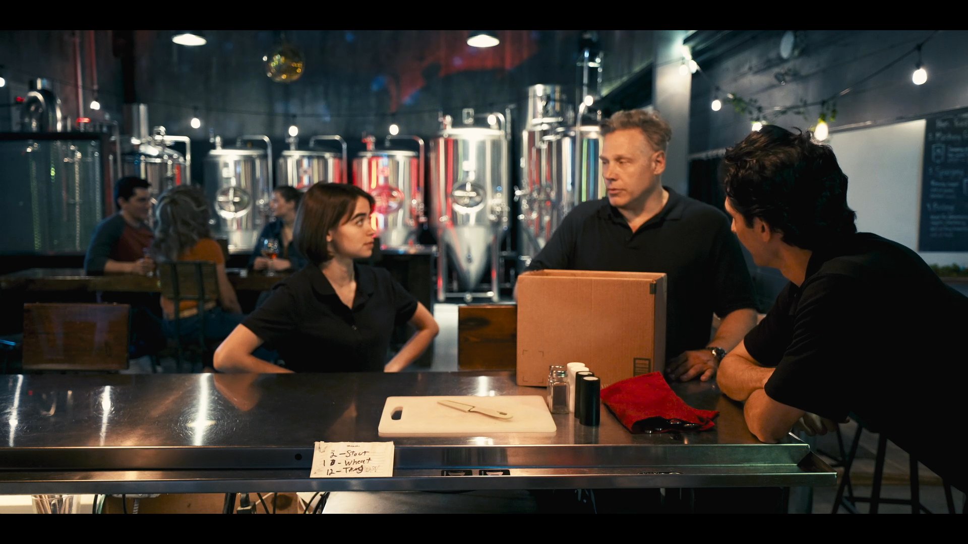 Two individuals engage in a focused professional discussion at a brewery counter, accompanied by a third colleague, while evaluating business materials. The setting suggests a collaborative project or operational planning session within an active brewery environment.