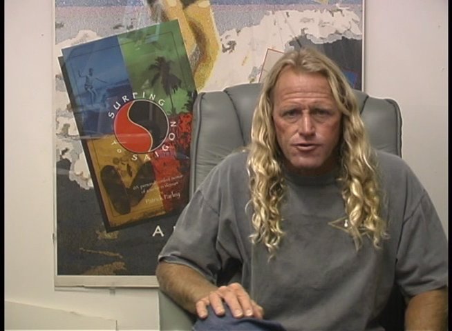 An experienced surfer discusses technical aspects of the sport during an interview session. He uses hand gestures to emphasize points while sitting in front of a decorative display.