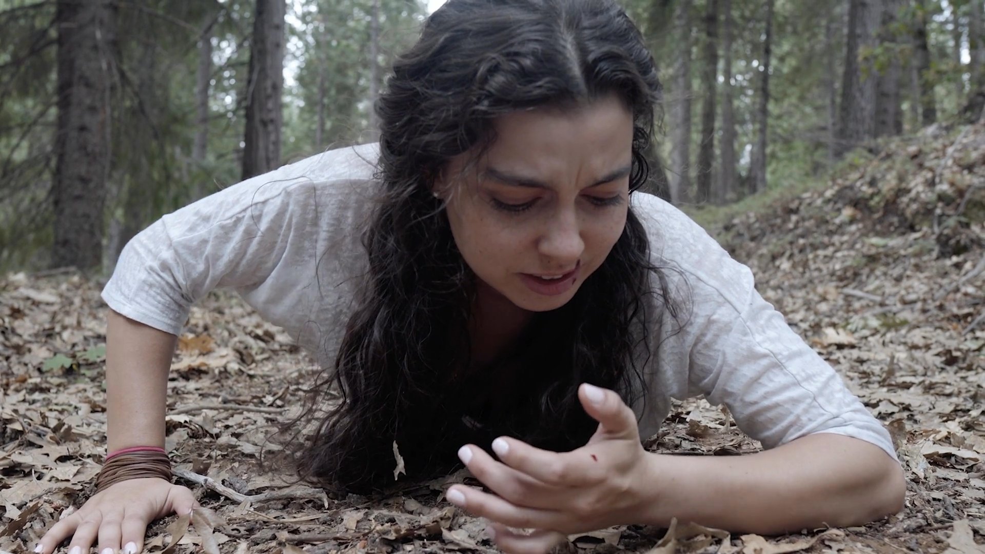 An injured hiker struggling through a forest landscape is monitored and approached by a drone. The aircraft captures close-up footage of the subject in distress on the forest floor.