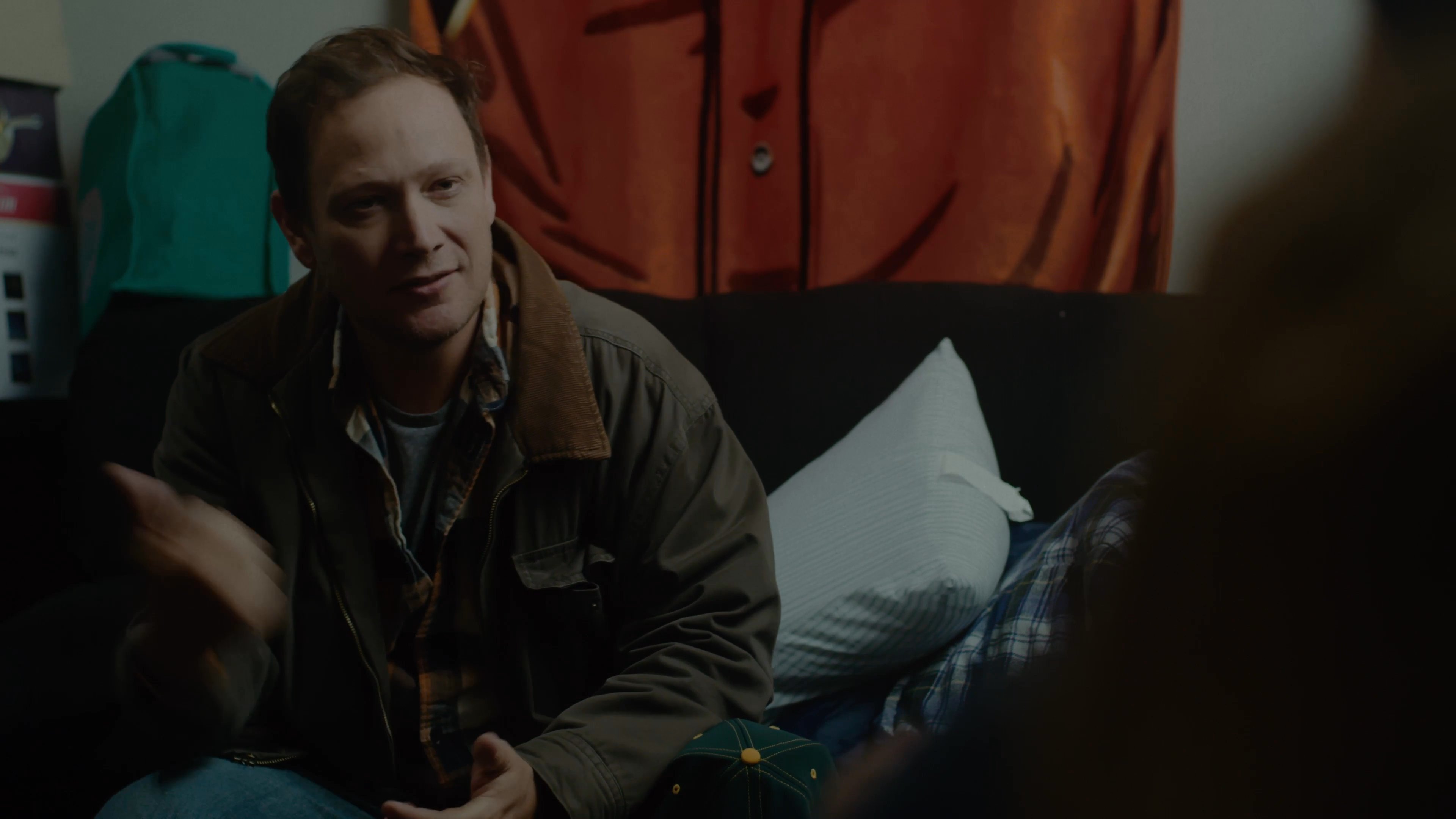 Two men sit in a living room and engage in a relaxed, seated conversation. One man eats from a small container while participating in the discussion.