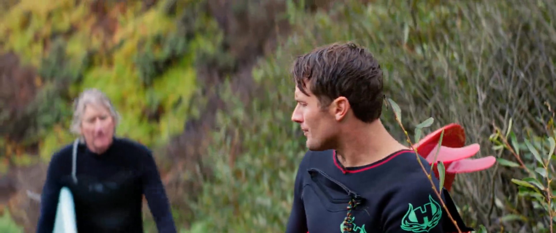 Two surfers dressed in wetsuits and carrying surfboards walk along a coastal path while engaged in conversation.