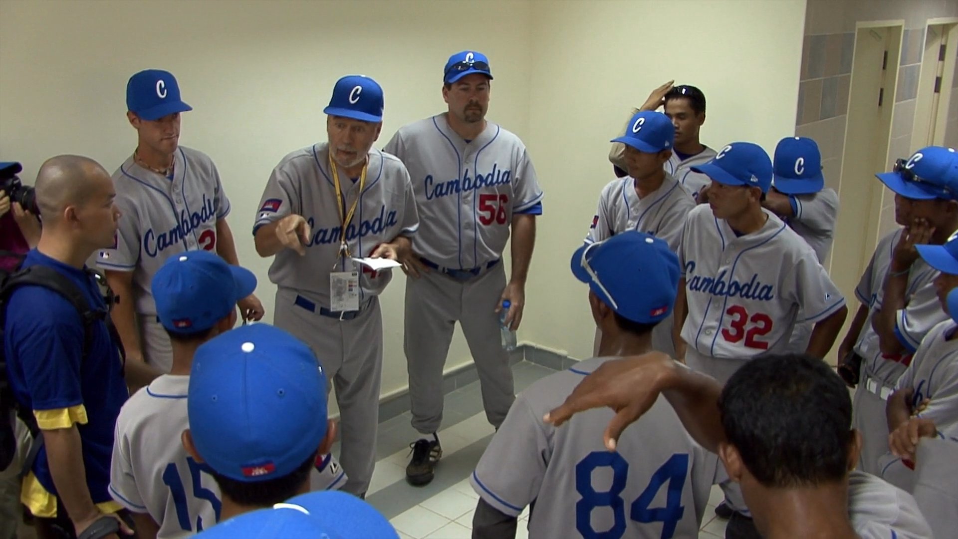 A baseball team in uniform gathers in a locker room to receive tactical instructions from their coaching staff. The players listen attentively as their coach gestures and discusses the game plan before taking the field.