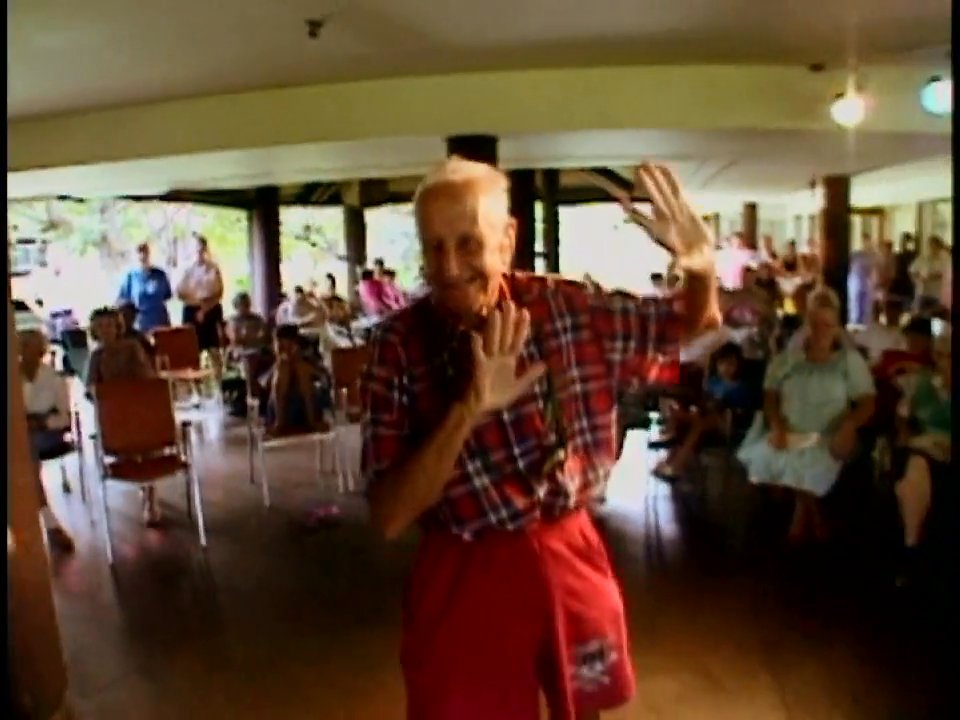 An elderly man performs a traditional hula dance for an audience gathered in a community space. His graceful movements demonstrate active participation in social and recreational programming.
