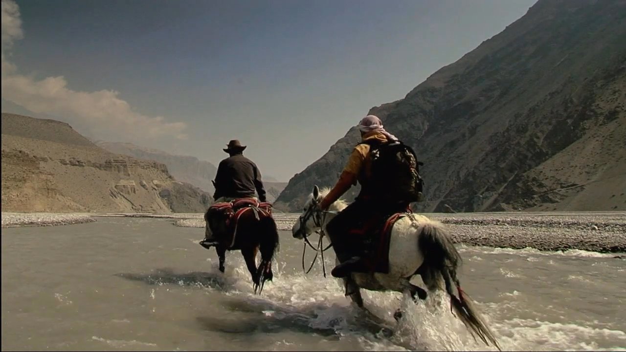 A group of travelers on horseback traverses a river and rocky terrain in the Mustang region of Nepal, passing ancient stupas and vibrant prayer flags.