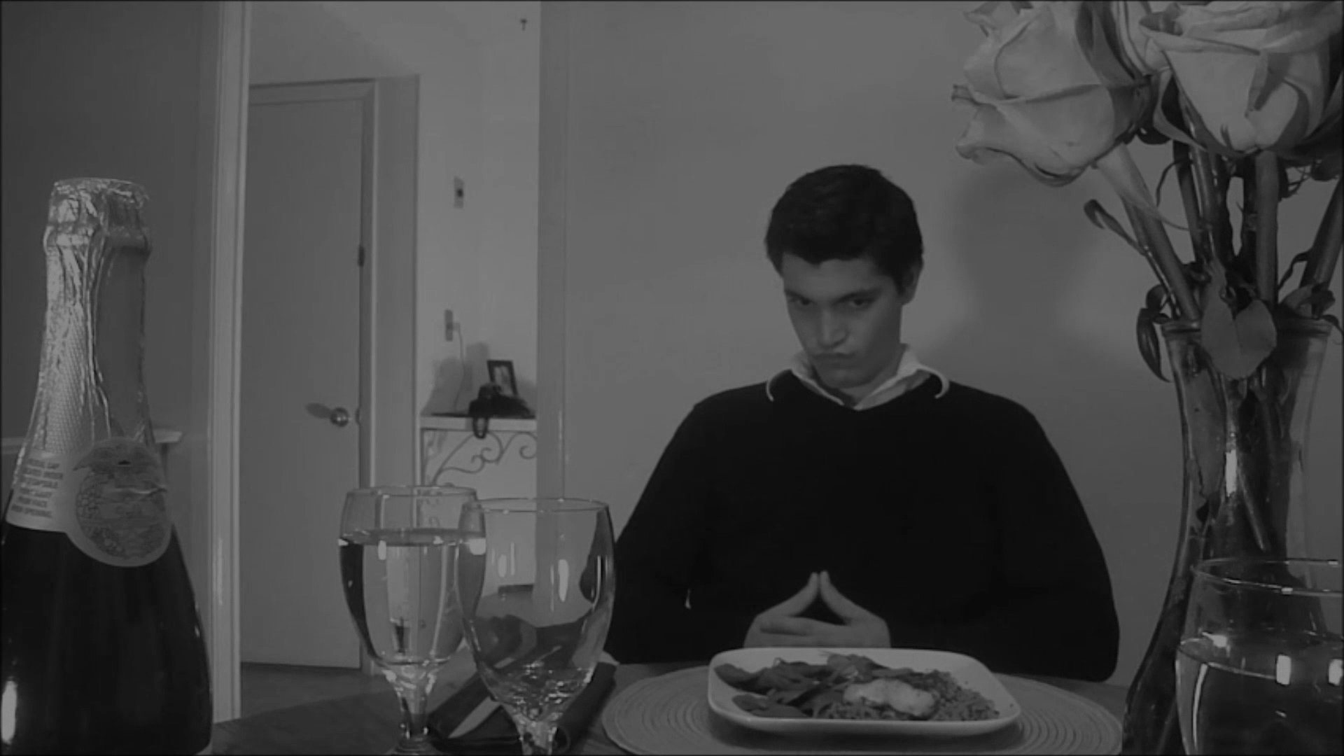 A man sits alone at a dining table set with a plate and a bottle of champagne, gradually shifting from an expectant expression to a somber, reflective mood. The atmosphere emphasizes themes of solitude and disappointment during a formal setting.