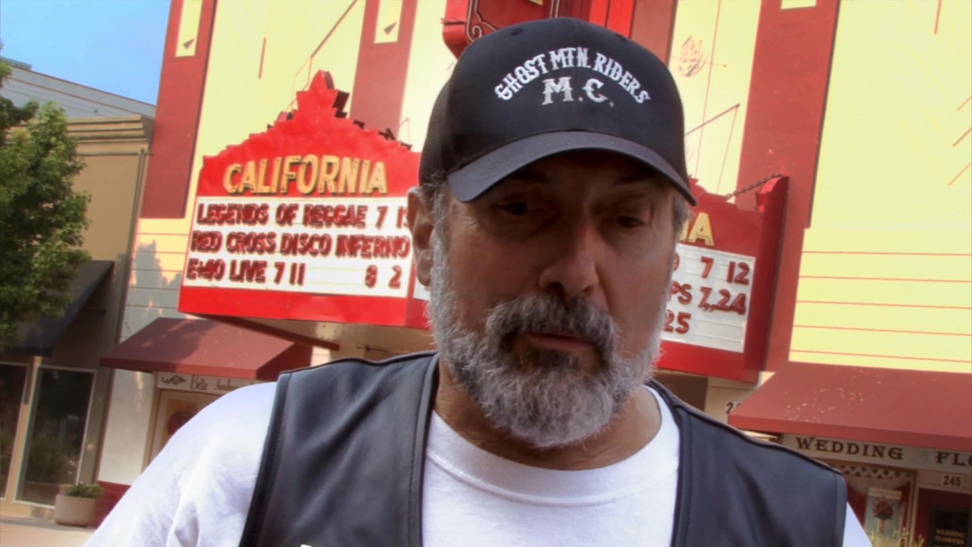 A man wearing a motorcycle club cap and vest provides an interview in front of a historic theater, followed by footage of a heavy rescue vehicle and a helicopter in the area.