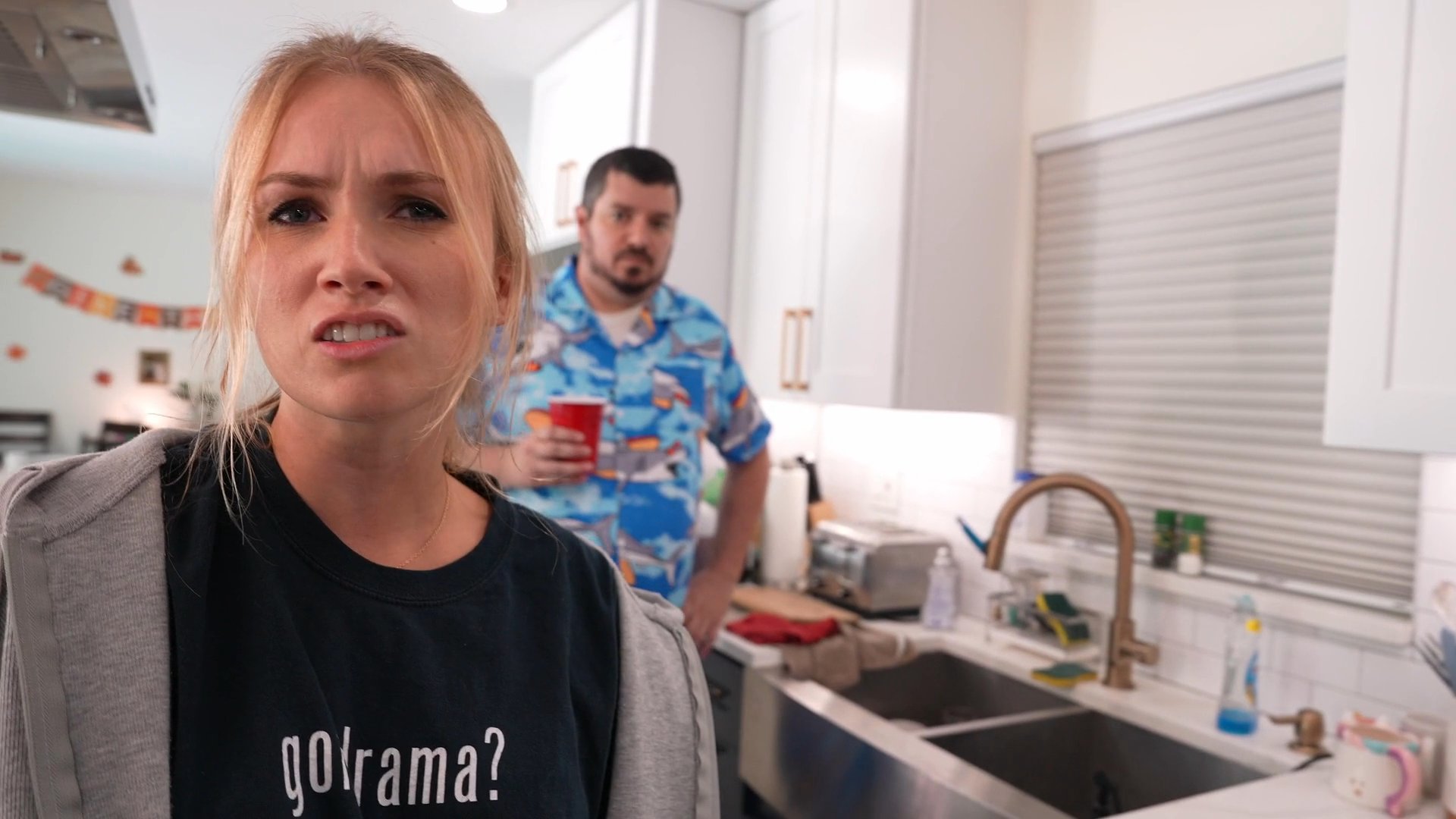 Two women argue in a kitchen as a man in a patterned shirt attempts to intervene in their tense disagreement.