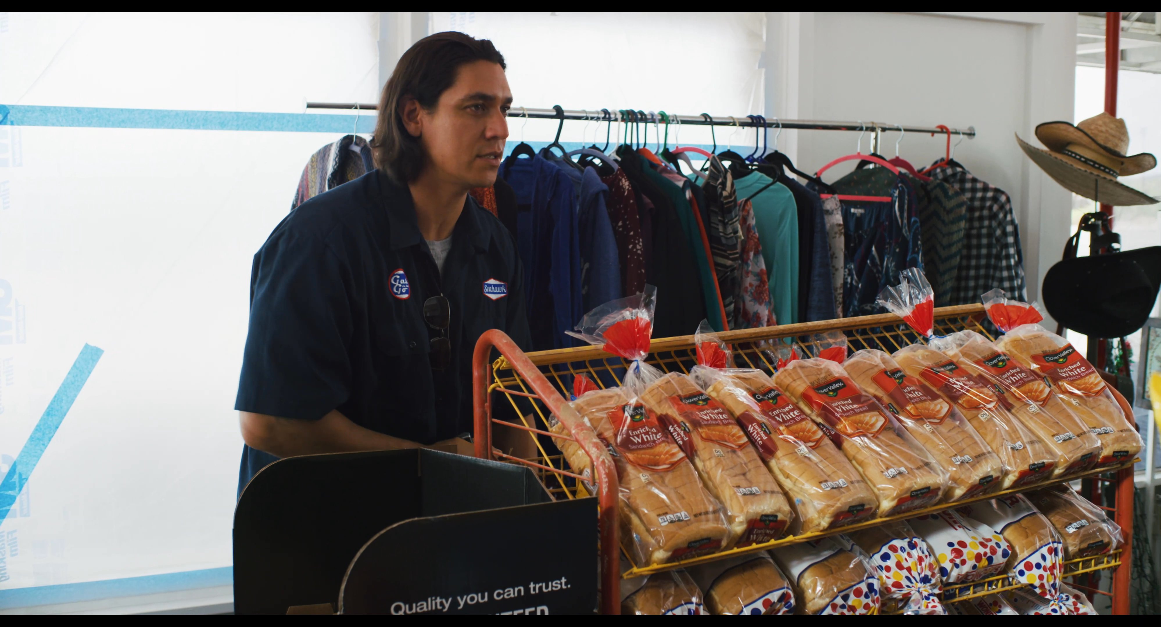 A customer and a store employee engage in a conversation inside a small retail shop. The individuals exchange dialogue while positioned near shelves stocked with packaged food items.