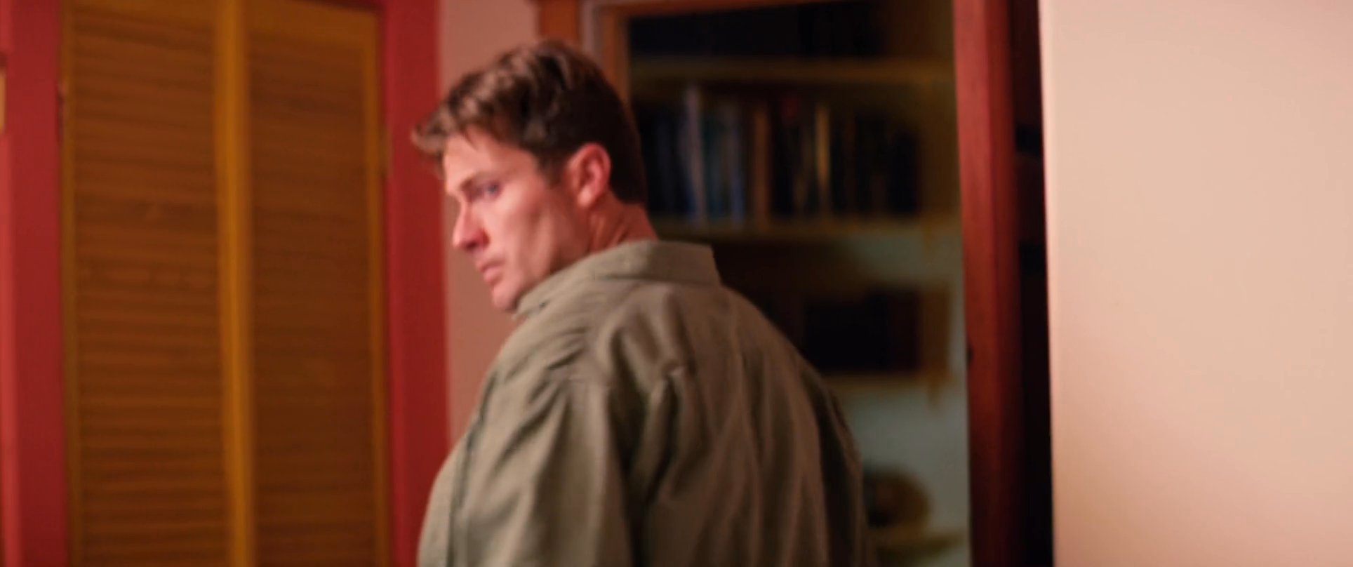 A man in casual clothing moves through an indoor space before settling by a window to converse. The scene shifts between his initial preparations and a focused, seated interaction.