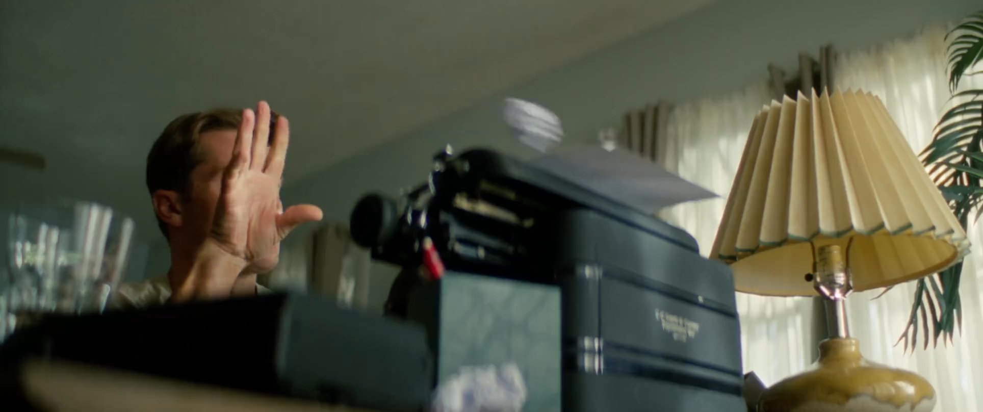 A man experiences creative frustration while working at a vintage typewriter, prompted by a framed photograph of surfers. He leans back in contemplation, surrounded by discarded paper, as he looks upward in thought.