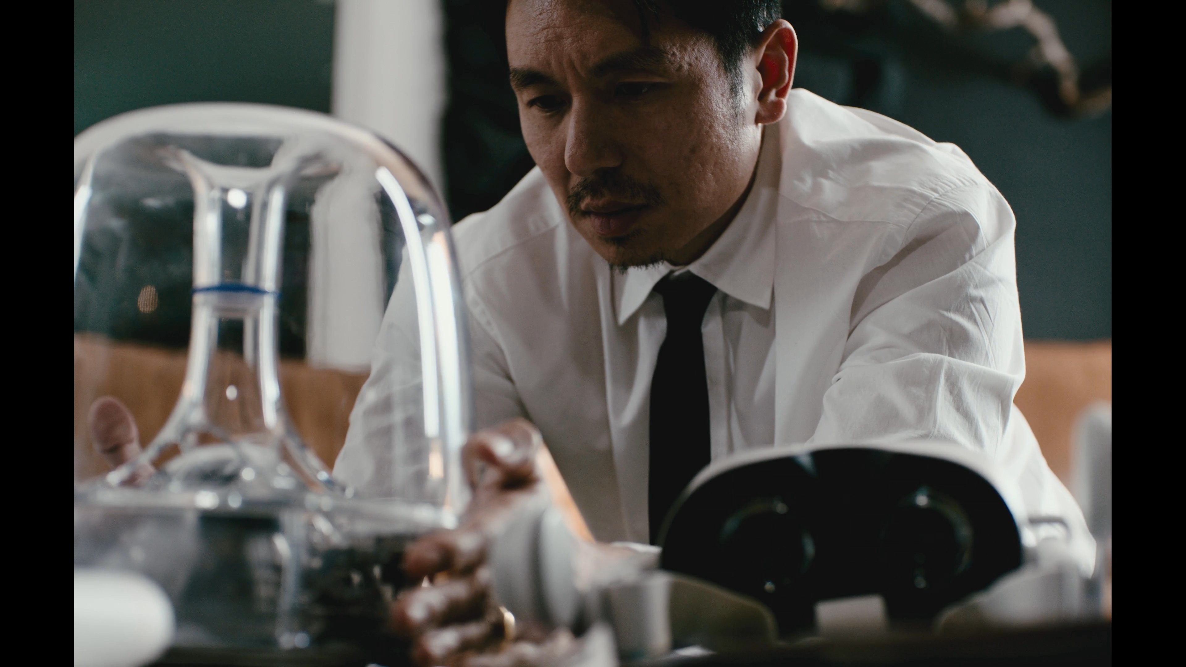 A woman with vibrant blue hair interacts with a man in a formal white shirt as he meticulously conducts a scientific experiment involving specialized glassware and equipment. The scene transitions between their focused expressions and the technical manipulation of mysterious laboratory apparatus.