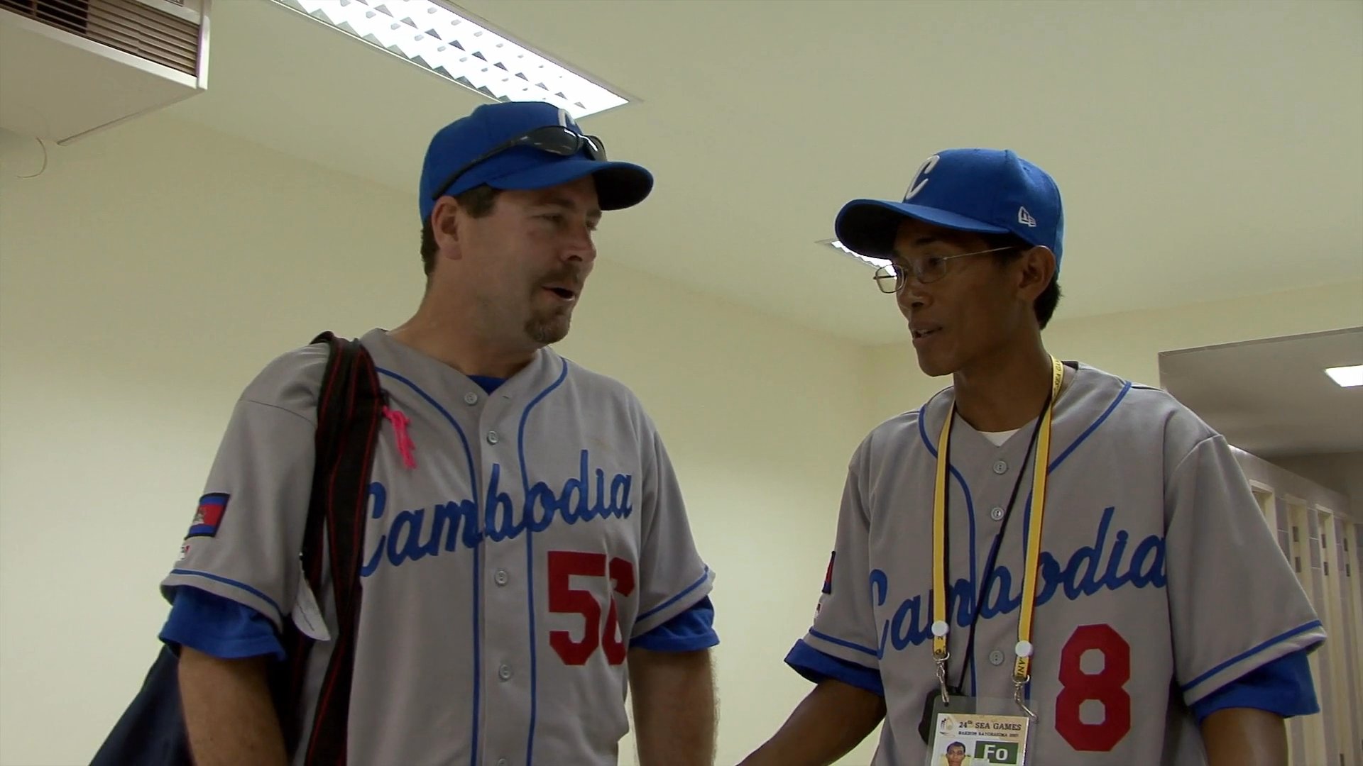 Two individuals in Cambodian baseball uniforms engage in a professional discussion before moving to an outdoor stadium to provide insights on the field. The interaction highlights the collaborative coaching dynamic and team preparation.