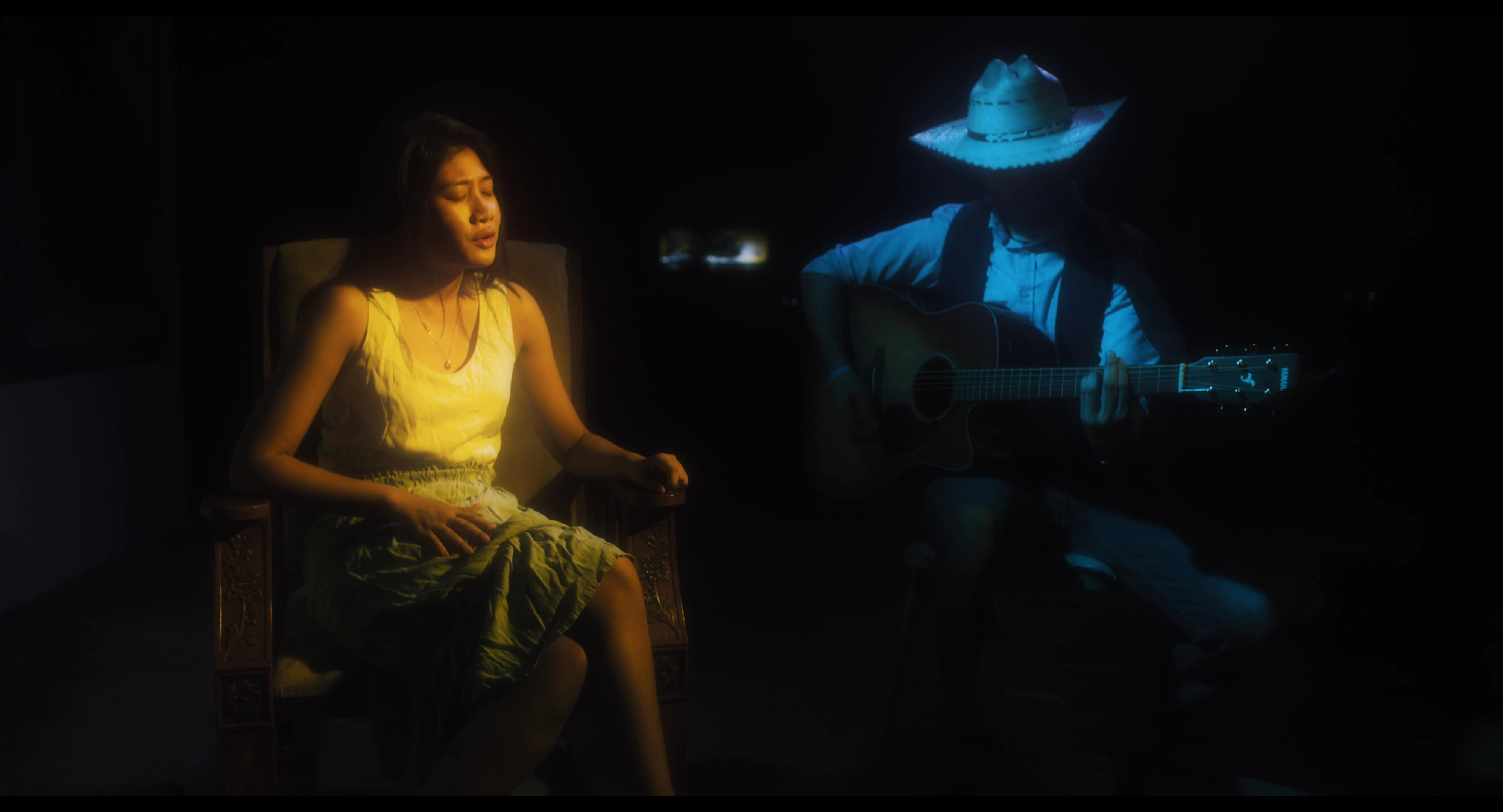 A man in a mechanic's uniform sits in a low-lit area before transitioning to a dimly lit room where a woman sings while accompanied by a guitarist wearing a cowboy hat.