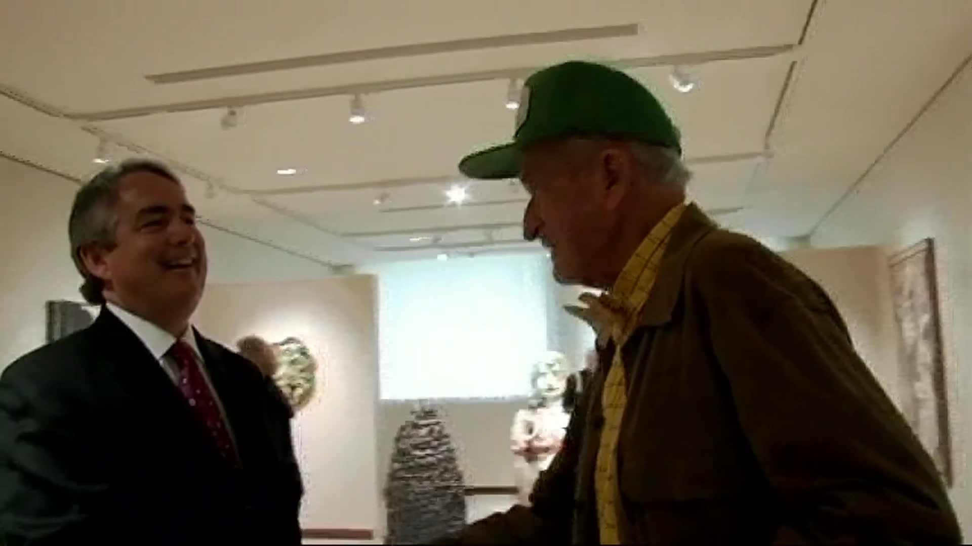 An older man in a green cap chats with a woman in a colorful jacket inside an art gallery. A suited man praises him, then they shake hands and embrace.
