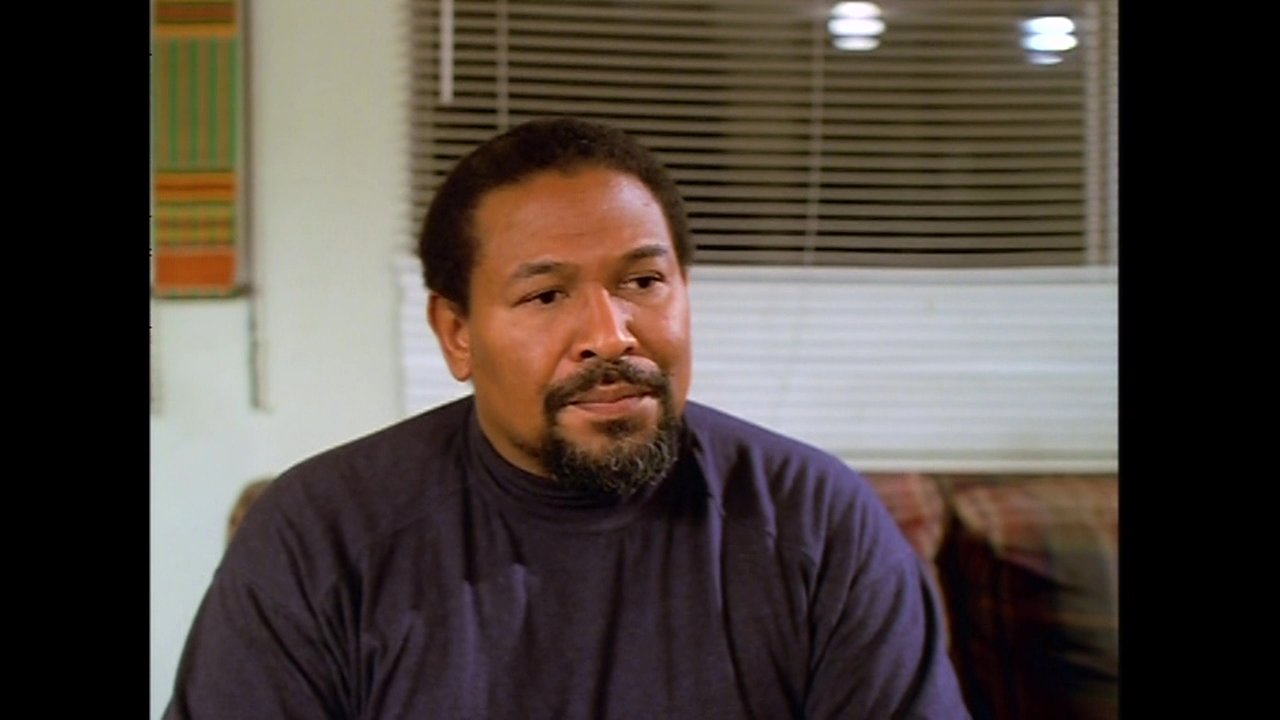 Two men engage in a serious, focused conversation within a residential living room setting. The participants exchange perspectives through active dialogue and direct interaction.