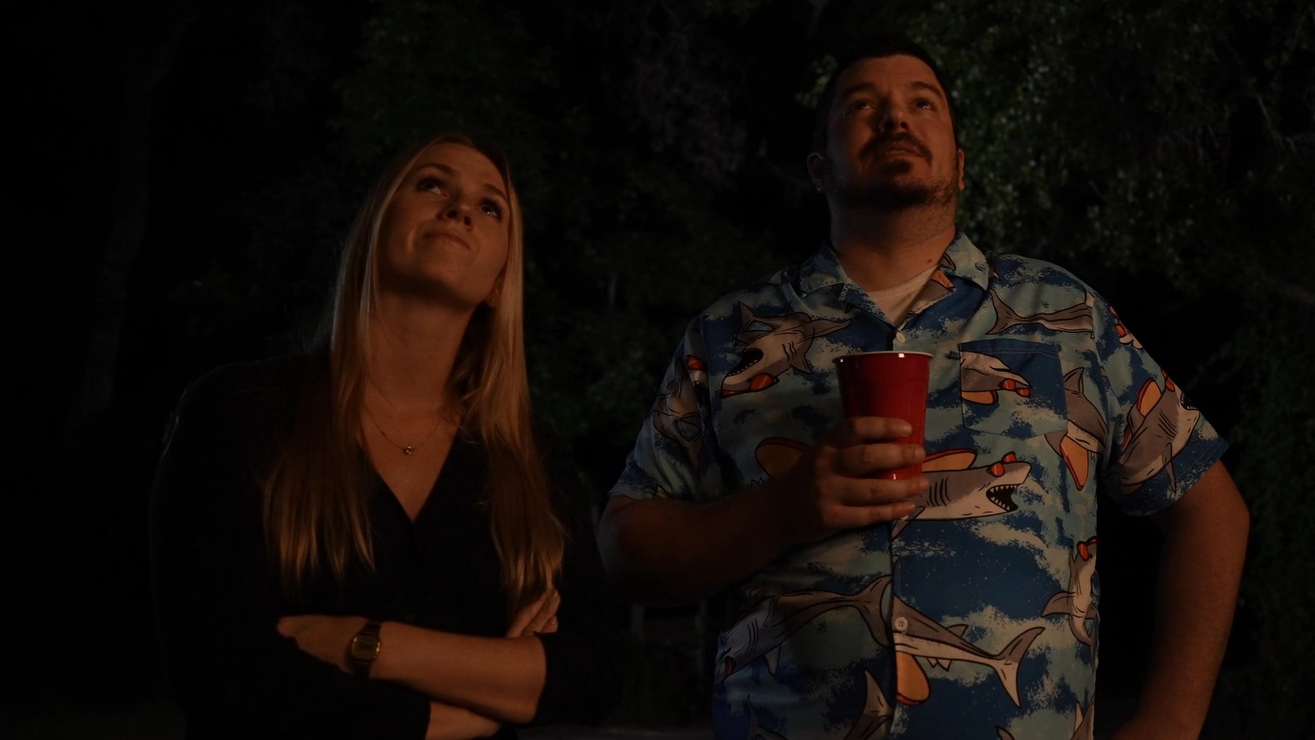 A man and a woman stand together at night, both looking upward intently. The man holds a drink while they observe the sky in a dark, outdoor setting.