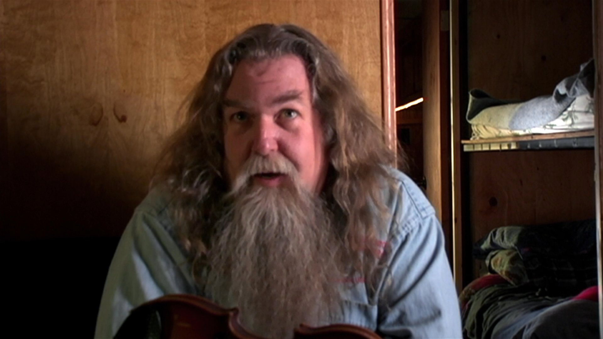 A long-haired, bearded man shares a personal story with expressive gestures while seated in a wood-paneled room. The scene concludes with a brief view through a window onto a desert road.
