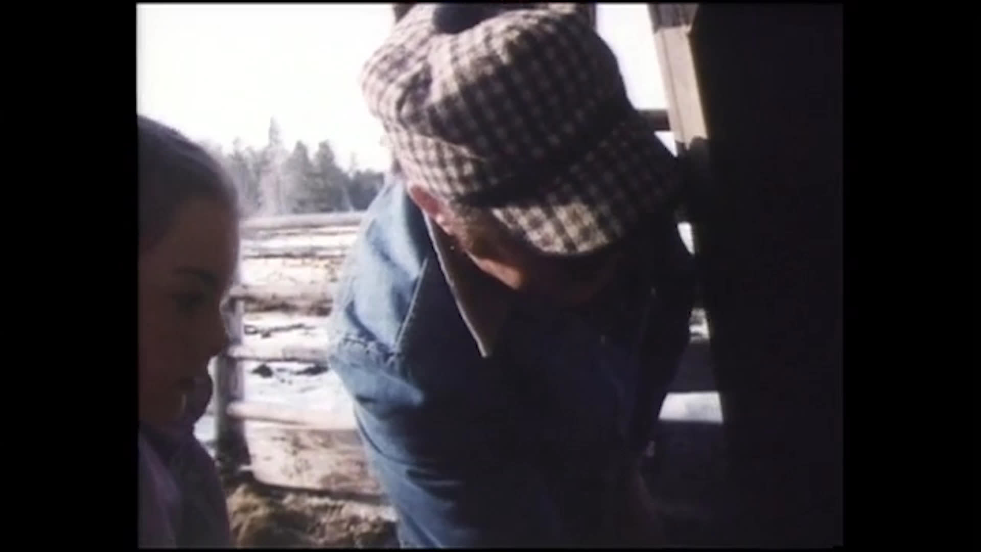 Two adults and a child work beside stacked hay bales on a snowy farm, then gather around a livestock chute. A cow stands in the chute as the adults attend to the equipment and discuss the process.