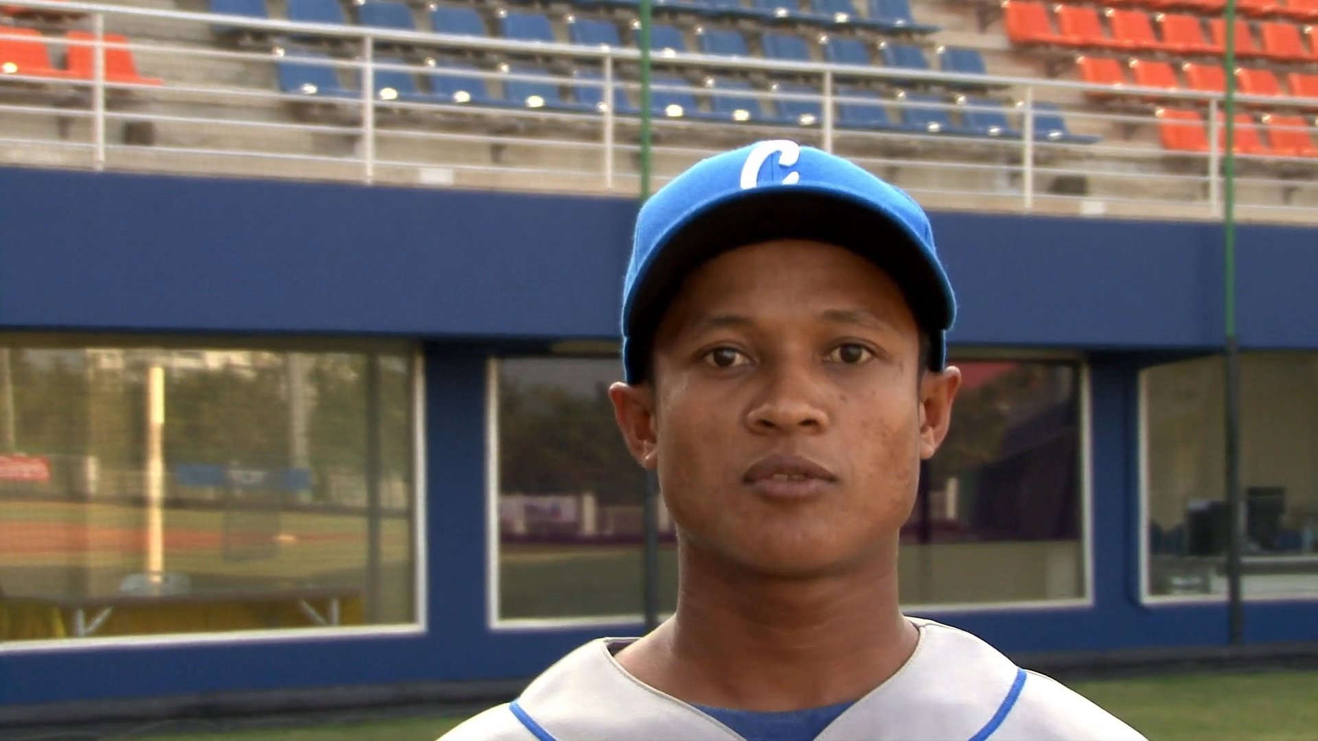 Baseball players and coaches stand on a professional field while sharing their experiences and perspectives in a series of interview segments. The participants are wearing team uniforms with the word Cambodia visible, reflecting their national representation.