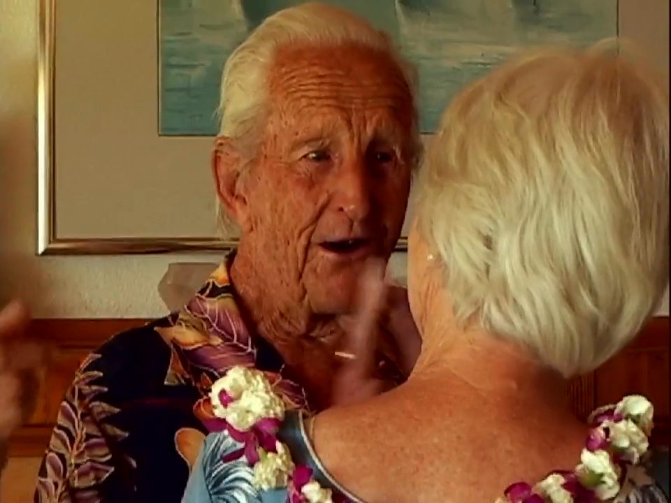 An elderly couple in tropical attire share an emotional embrace and engage in heartfelt conversation within a home environment.