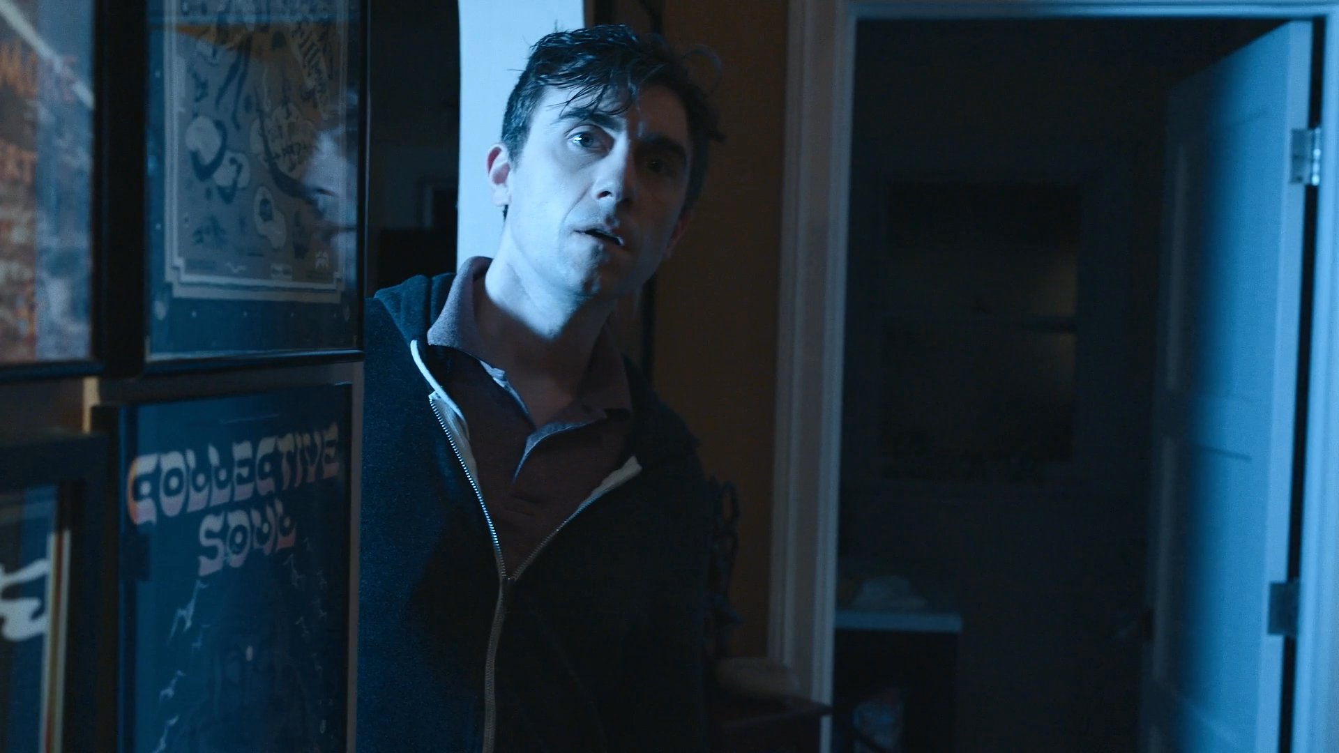 A man moves cautiously through a residential interior to observe an elderly woman working in the kitchen. The scene captures an atmosphere of quiet surveillance within a home environment.