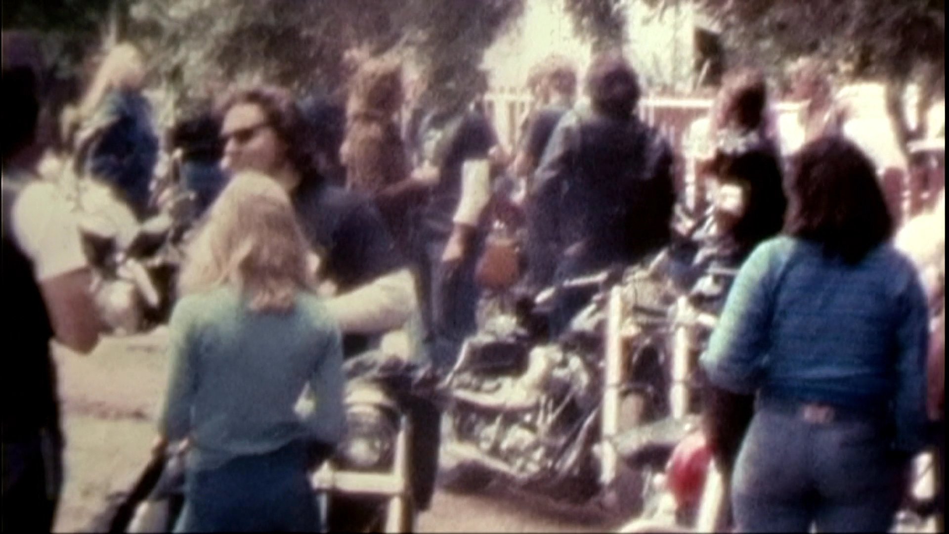 The footage transitions from archival shots of a large motorcycle club gathering in a natural setting to a contemporary interview with an individual sharing insights on the subculture. The sequence contrasts the candid, energetic atmosphere of a historical biker event with a reflective, personal account of that era.