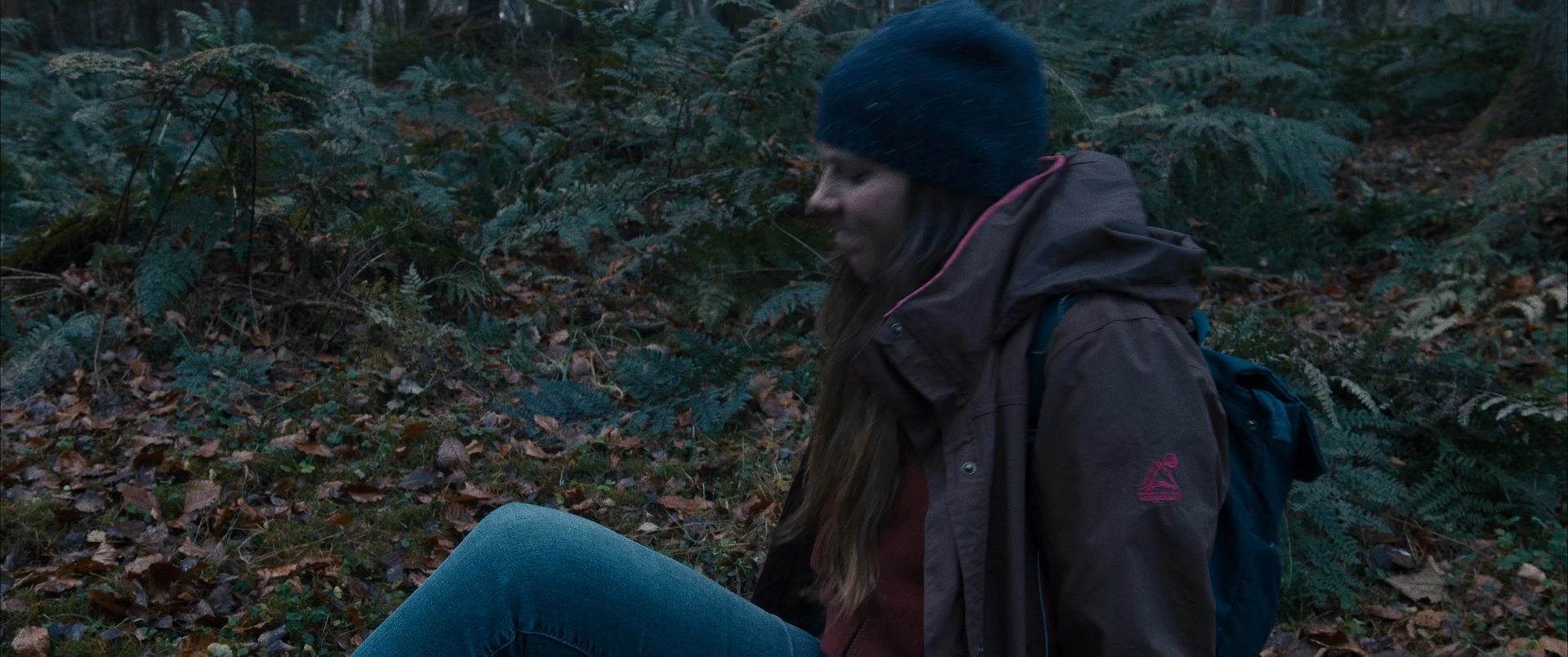 A hiker resting in a forest receives help getting back on her feet from a passerby. The two individuals engage in a brief, supportive exchange within the woodland environment.